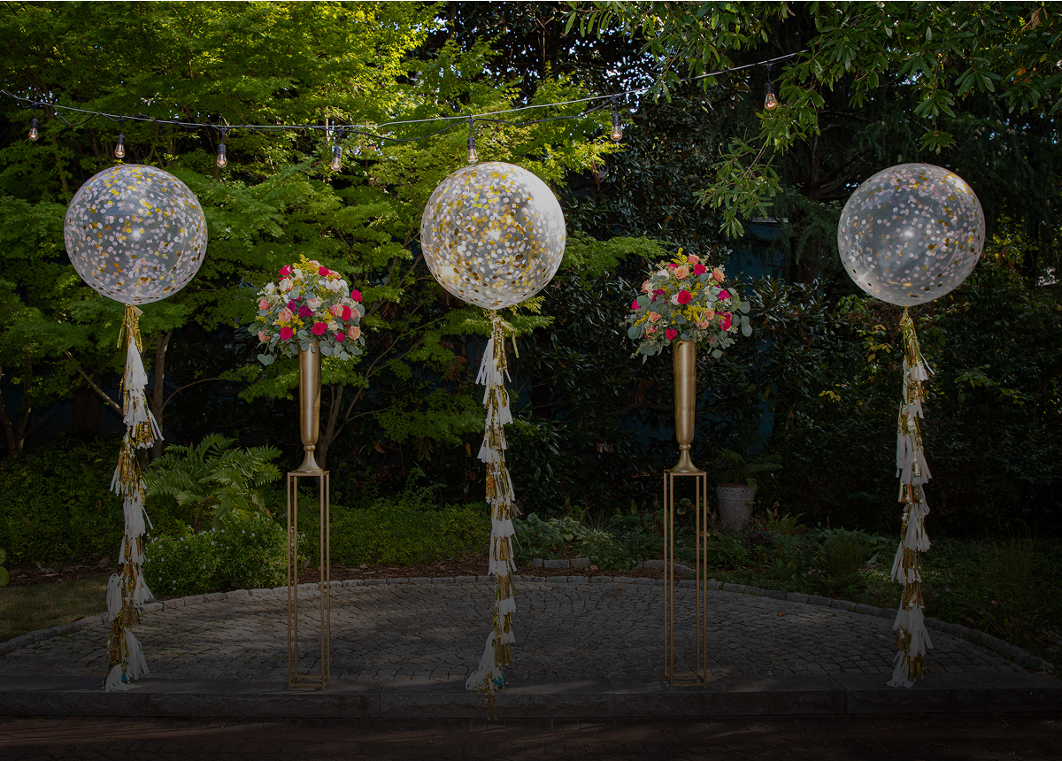 Four tall gold stands hold two confetti-filled balloons and two flower arrangements with pink and orange flowers, set outdoors with greenery in the background.