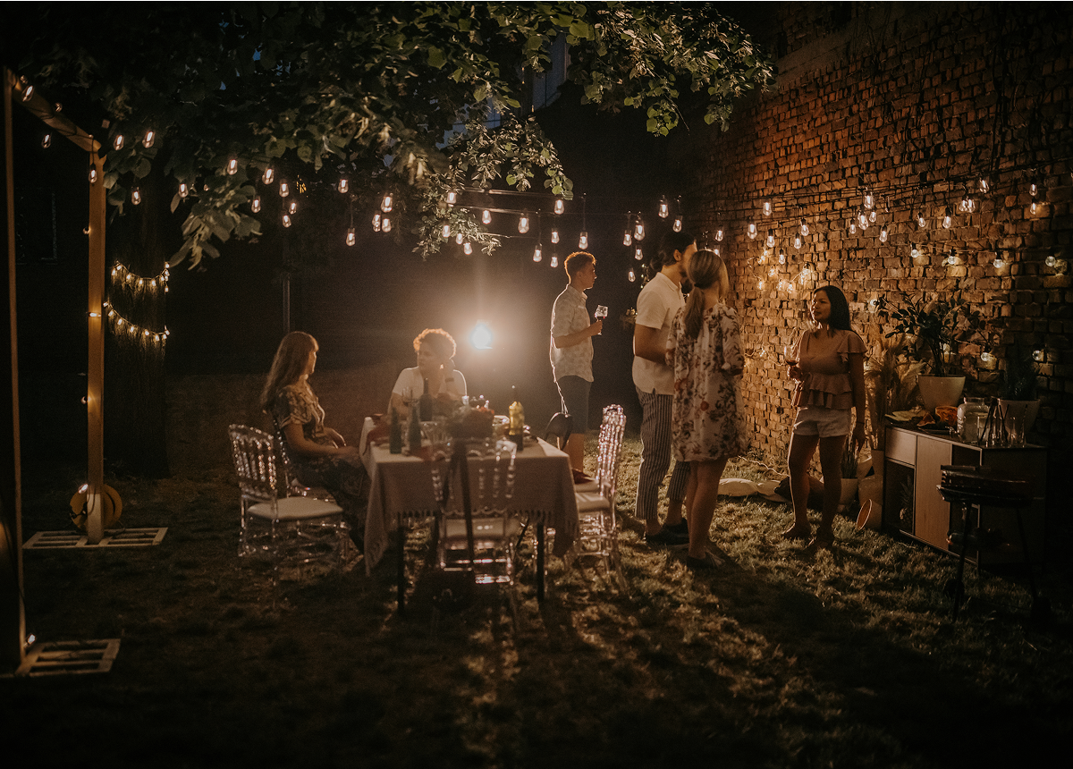 A group of people socialize and dine in a backyard at night, surrounded by string lights and a brick wall, creating a cozy outdoor atmosphere.