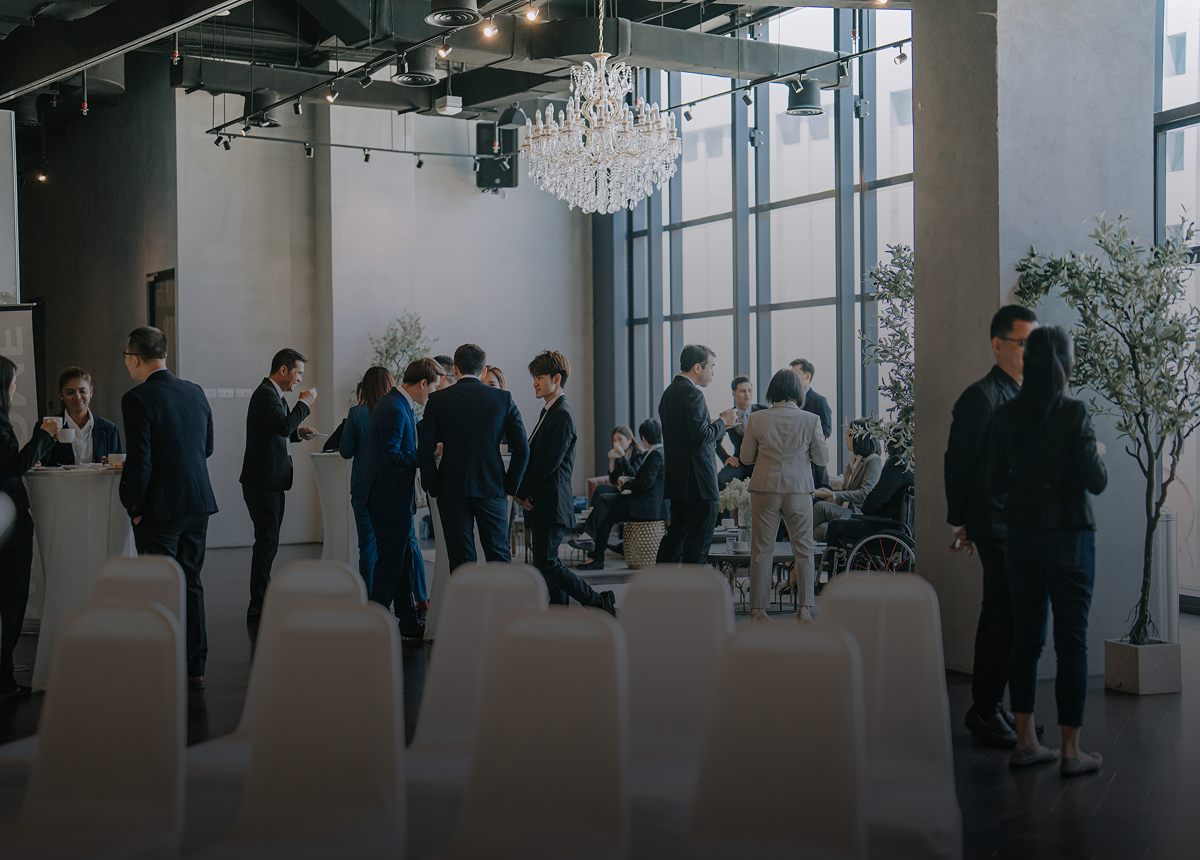 A group of people in business attire are gathered and conversing in a modern, well-lit event space with large windows, white chairs, and a chandelier.